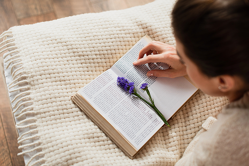 high angle view of blurred young woman reading book with purple flower while resting on bed at home
