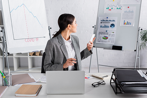 Young businesswoman with smartphone looking at flipchart near documents and laptop in office