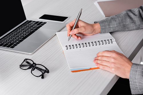 Cropped view of businesswoman writing on notebook near eyeglasses and blurred gadgets