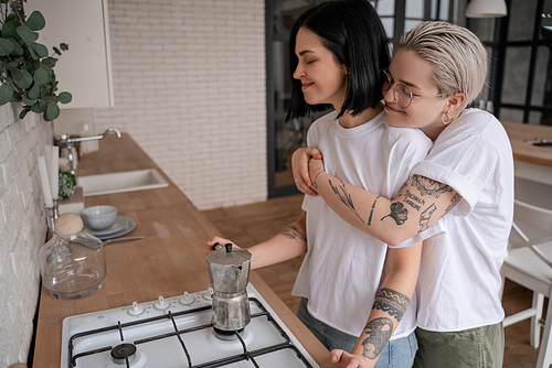 young woman in glasses hugging happy girlfriend near coffee pot on stove