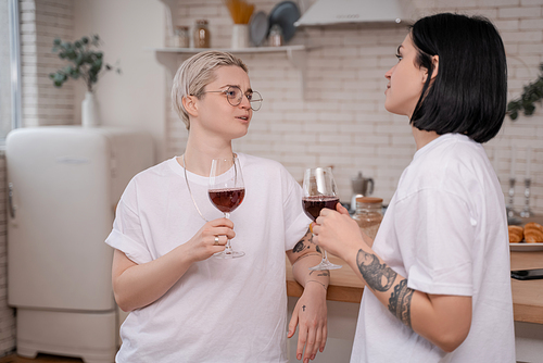 young lesbian couple holding glasses of red wine while talking in kitchen