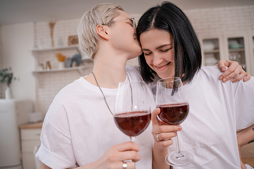 woman in glasses kissing happy girlfriend while holding glasses of red wine