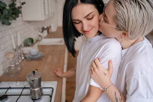 young woman in glasses kissing cheek of girlfriend near coffee pot on blurred background