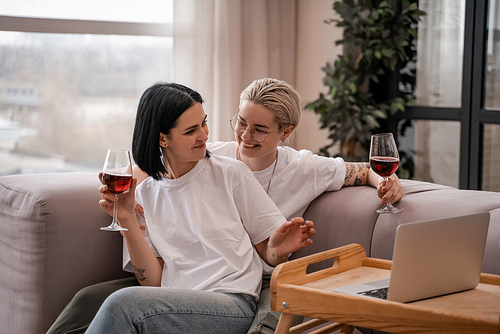 happy lesbian couple watching movie on laptop while holding glasses of red wine