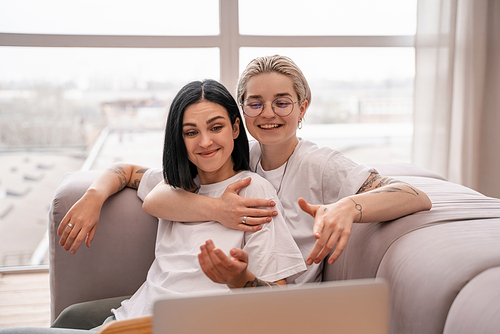 young lesbian couple smiling while chilling on sofa near laptop