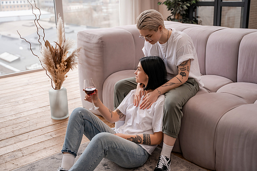tattooed woman looking at happy girlfriend with glass of red wine