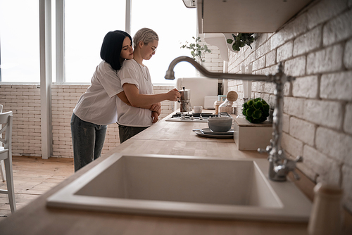 young woman hugging girlfriend near coffee pot on stove with sink on blurred foreground