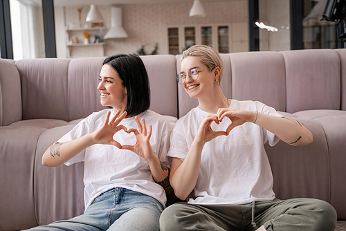 smiling lesbian couple showing heart sign with hands in living room