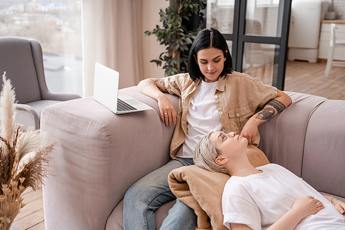 tattooed woman sitting on couch near laptop and looking at girlfriend