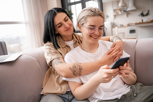 happy lesbian couple looking at smartphone while chilling on sofa