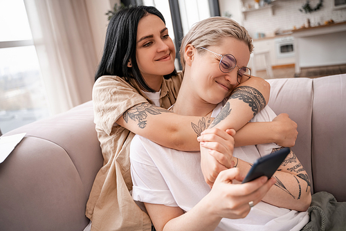 lesbian couple hugging while sitting on couch and using smartphone
