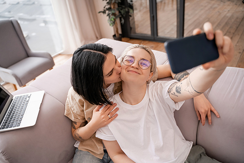 woman kissing cheek of happy girlfriend in glasses taking selfie in living room