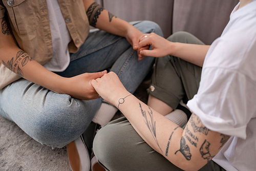 cropped view of tattooed lesbian couple sitting and holding hands in living room