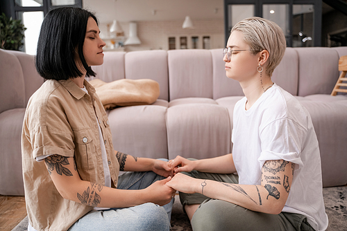 side view of lesbian couple sitting and holding hands while meditating in living room