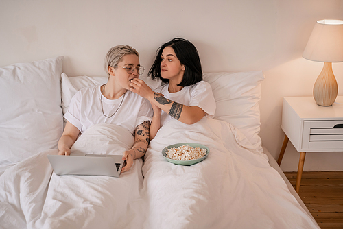 brunette woman feeding girlfriend with popcorn near laptop on bed