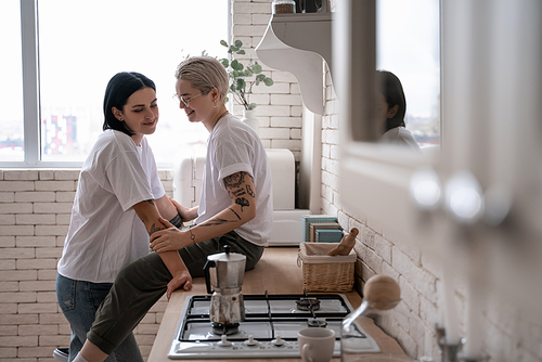 happy young woman sitting on kitchen table and hugging with girlfriend near coffee pot on stove