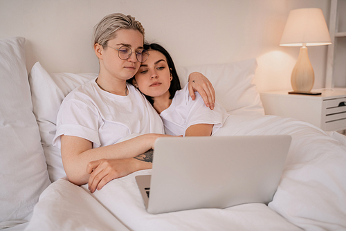 young lesbian couple hugging while lying on bed and watching movie on laptop