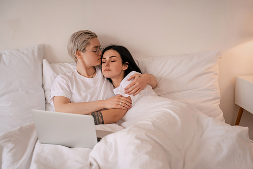 young lesbian couple hugging while lying on bed near laptop
