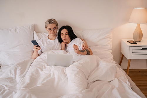 young lesbian couple lying in bed and watching movie on laptop