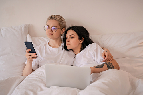 young lesbian couple lying in bed and looking at smartphone near laptop