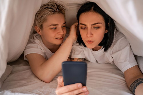young woman texting on smartphone while lying under blanket with girlfriend