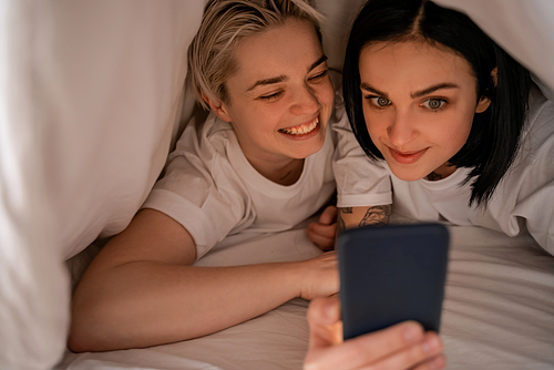 young woman messaging on smartphone while lying under blanket with girlfriend