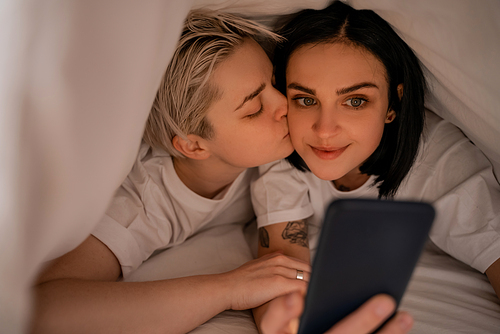 young woman lying under blanket and kissing girlfriend texting on smartphone