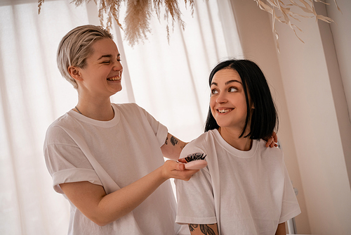 cheerful young woman brushing hair of brunette girlfriend in bedroom