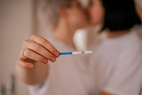 young lesbian woman holding pregnancy test and kissing girlfriend on blurred background