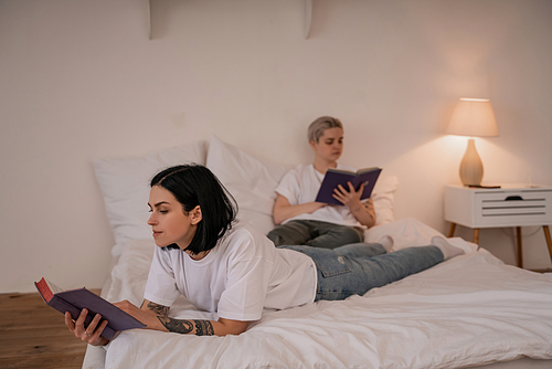 brunette woman lying on bed and reading book near girlfriend on blurred background