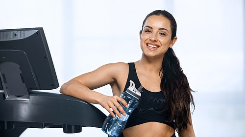 happy woman in sportswear holding sports bottle with water near treadmill