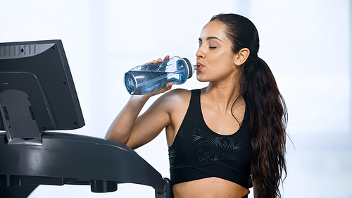 young woman in sportswear holding sports bottle and drinking water near treadmill