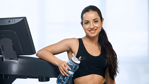 cheerful woman in sportswear holding sports bottle with water near treadmill