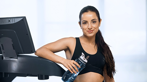 young pleased woman in sportswear holding sports bottle with water near treadmill