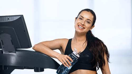joyful woman in sportswear holding sports bottle with water near treadmill