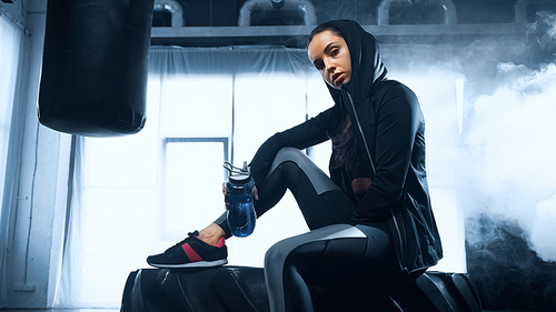 sporty woman in hoodie sitting on tire with sports bottle and 