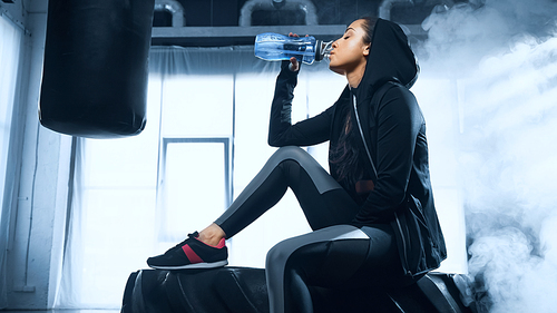 sportswoman in hoodie sitting on tire and drinking water from sports bottle