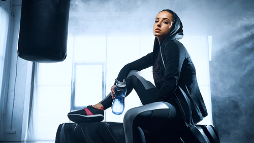 sportswoman in hoodie sitting on tire  with sports bottle and 