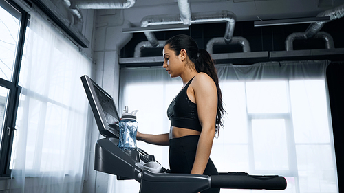 sweaty sportswoman using touchscreen on treadmill near sports bottle with water