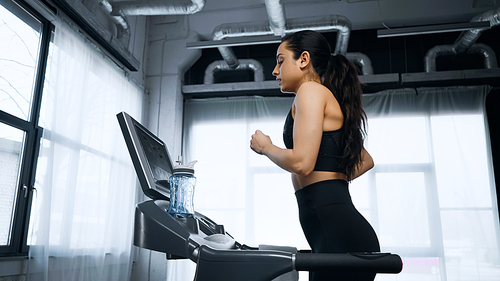 side view of sportswoman running on treadmill near sports bottle with water in gym