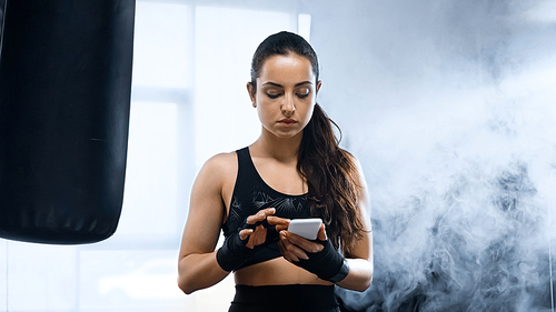 young brunette sportswoman with boxing bandages on hands using smartphone