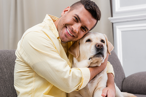 pleased man smiling at camera while cuddling labrador at home