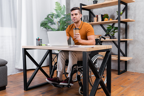 disabled man holding coffee to go while working near laptop at home