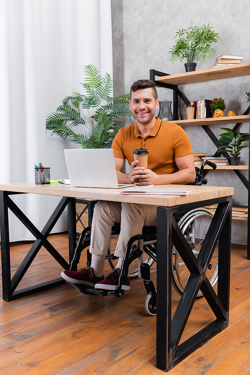 handicapped man smiling at camera while holding coffee to go near laptop