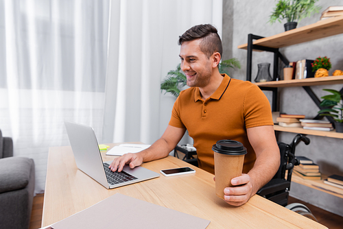positive, disabled man holding coffee to go while typing on laptop near cellphone with blank screen