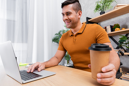 cheerful, disabled man holding coffee to go while typing on laptop at home