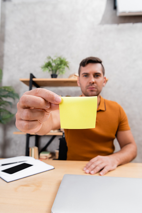 young man showing empty sticky note while working at home on blurred background