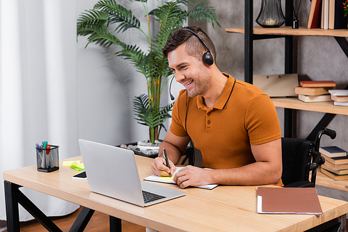 smiling, disabled man writing in notebook while working in headset at home