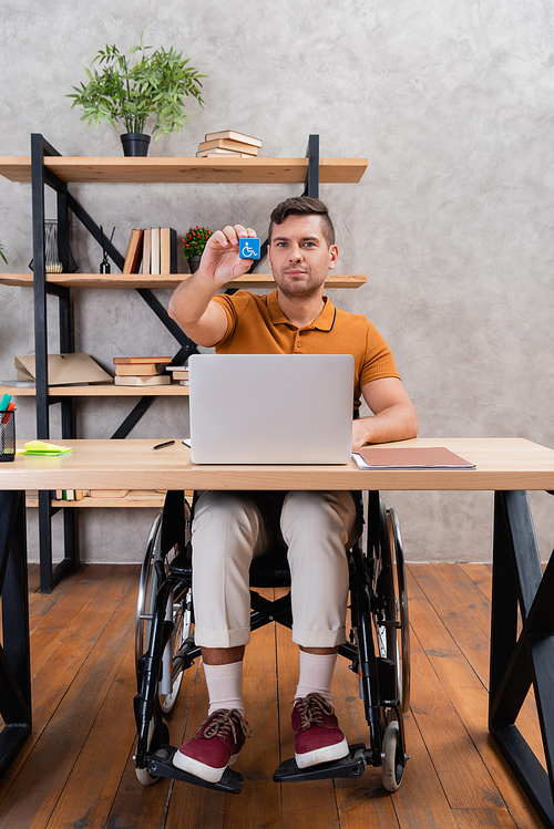 young man showing disability sign while sitting in wheelchair in home office