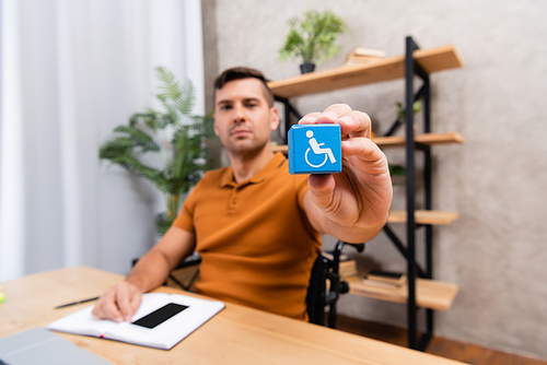 young man showing cube with disability sign while sitting in home office, blurred background